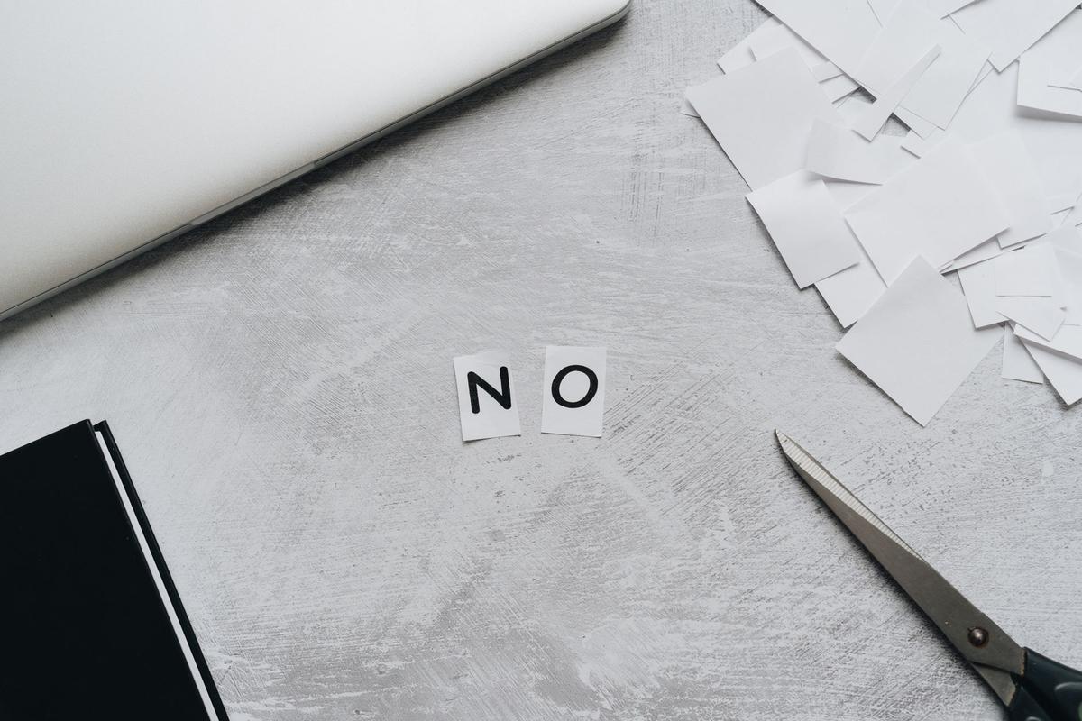 Top view of scattered paper squares, a laptop, and scissors arranged to spell the word 'NO' on a desk