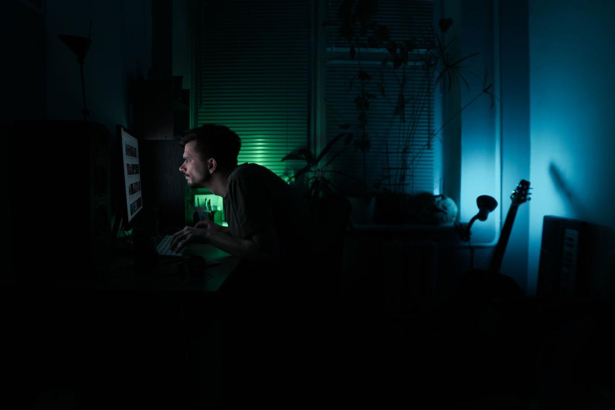Side view of a young bearded man working alone on a computer in a dark room at home