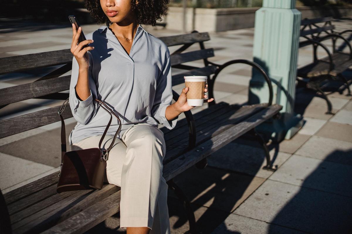 Relaxed woman sitting outdoors on a bench, holding a coffee and looking at her smartphone