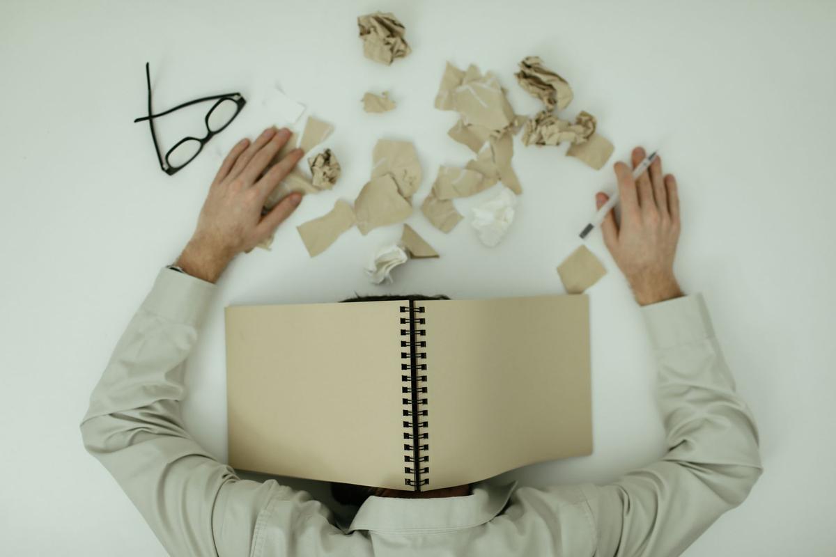 Overhead view of an exhausted man lying with a notepad over his head, surrounded by crumpled papers